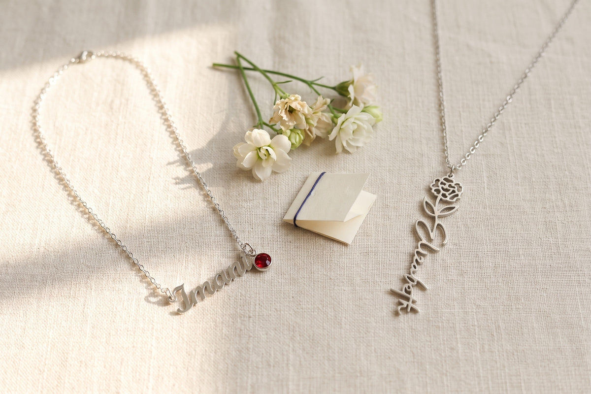 Silver name necklace with a red birthstone beside a birth-flower name pendant on linen