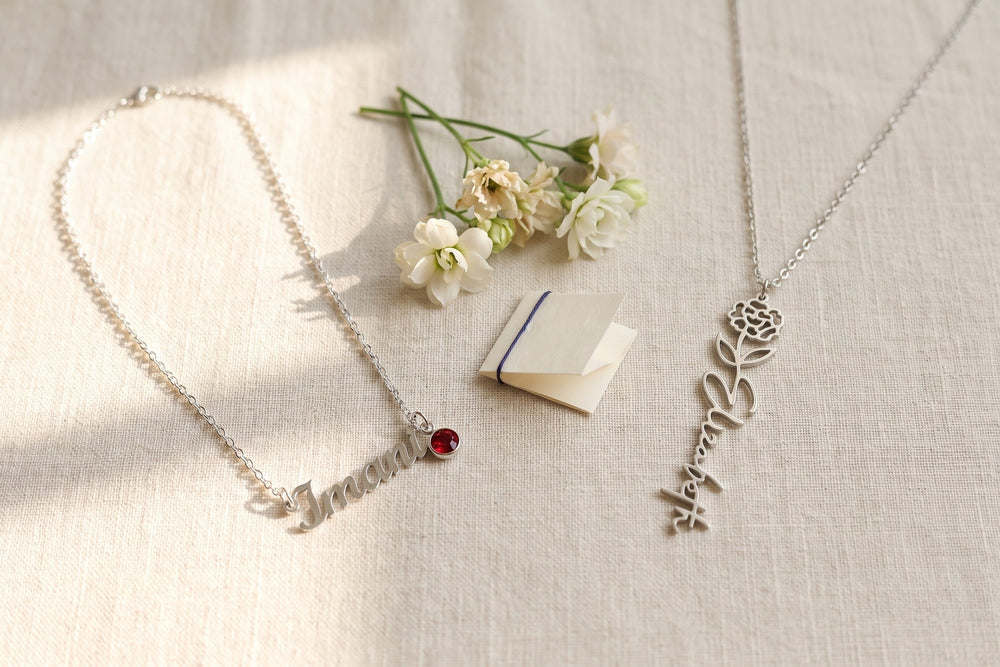 Silver name necklace with a red birthstone beside a birth-flower name pendant on linen