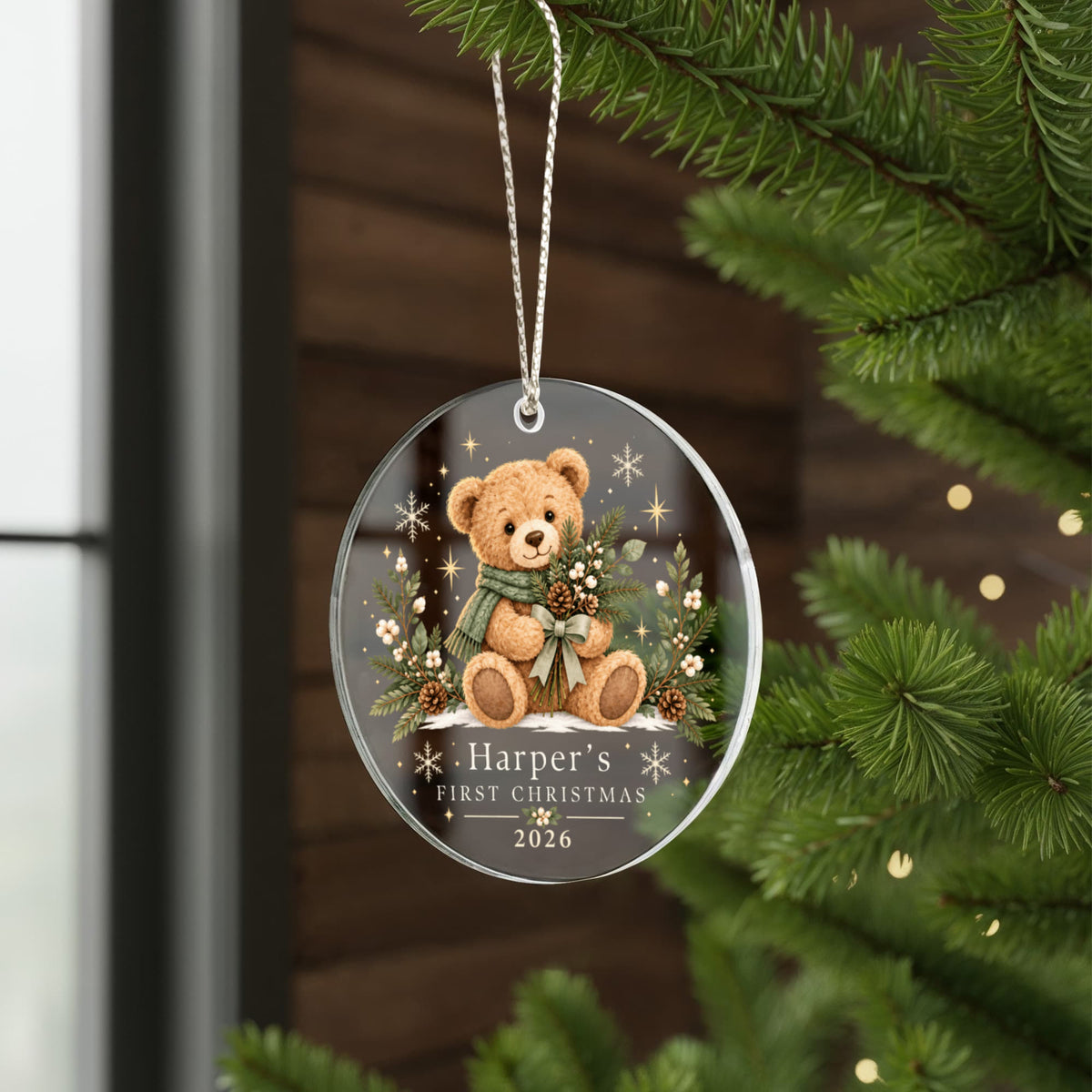Evergreen teddy first Christmas ornament hanging beside fir branches by a window.