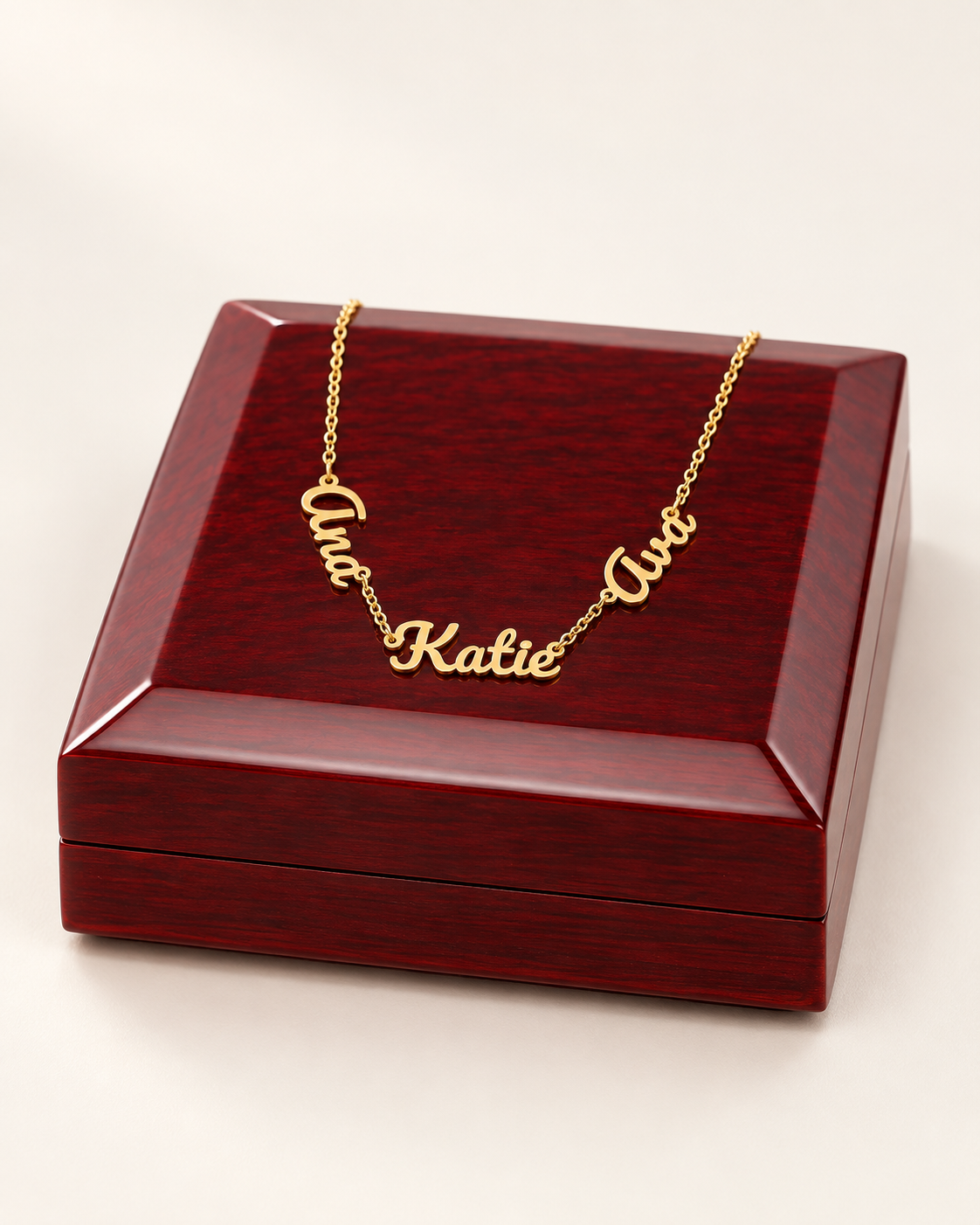 Gold-tone three-name necklace draped across a wooden gift box on ivory.