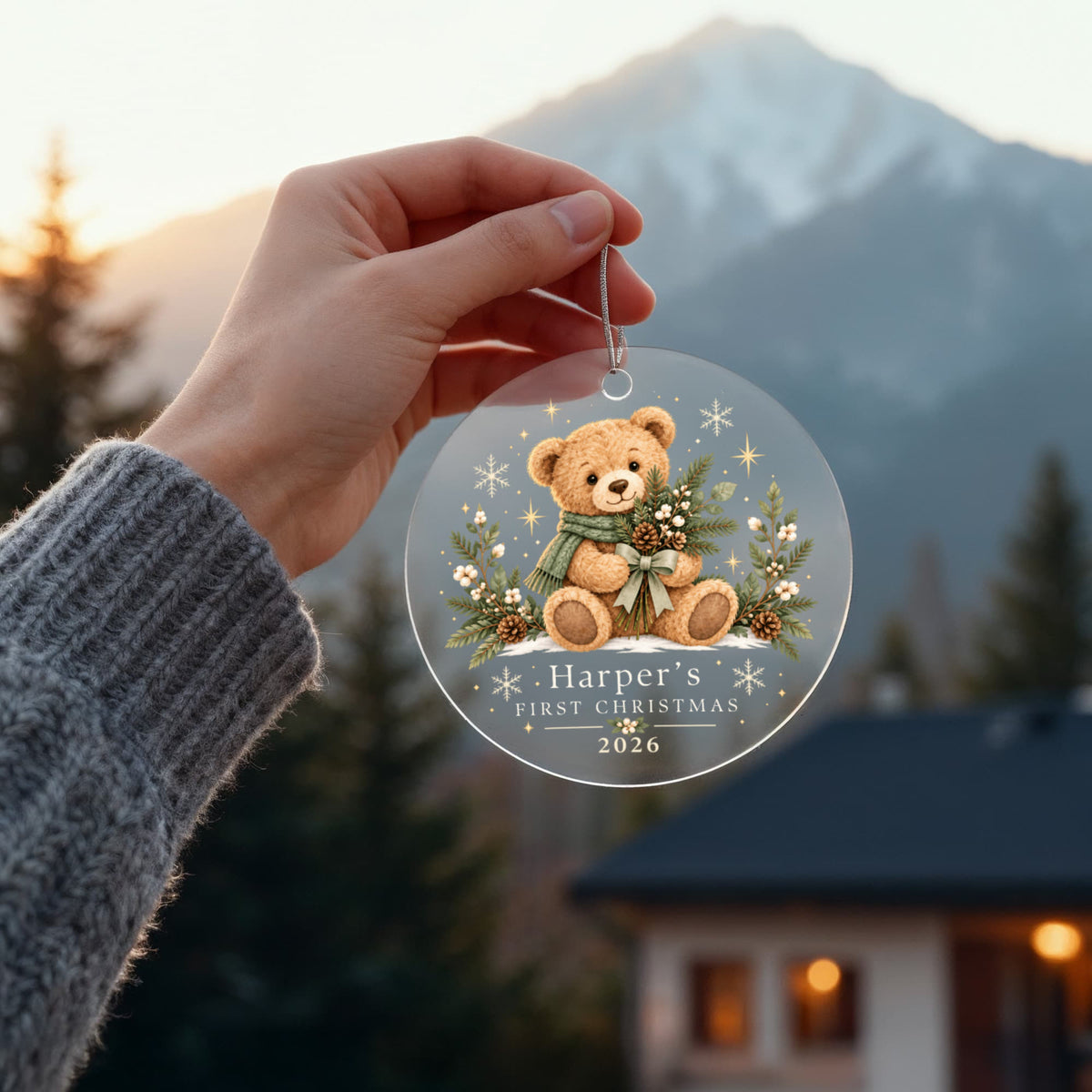 Hand holding an evergreen teddy first Christmas ornament outdoors with mountains in the background.