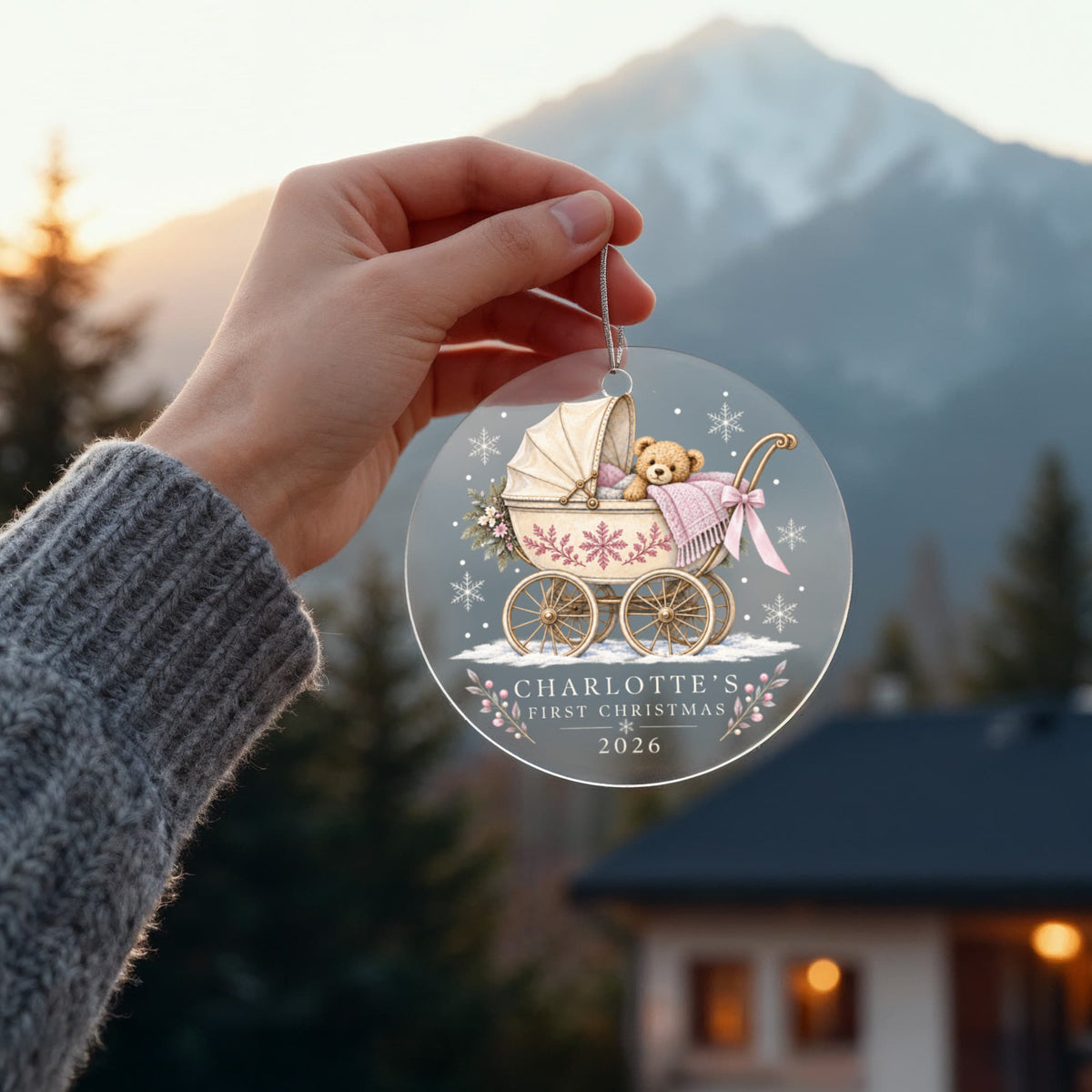 Hand holding a pink teddy pram first Christmas ornament outdoors with mountains in the background.