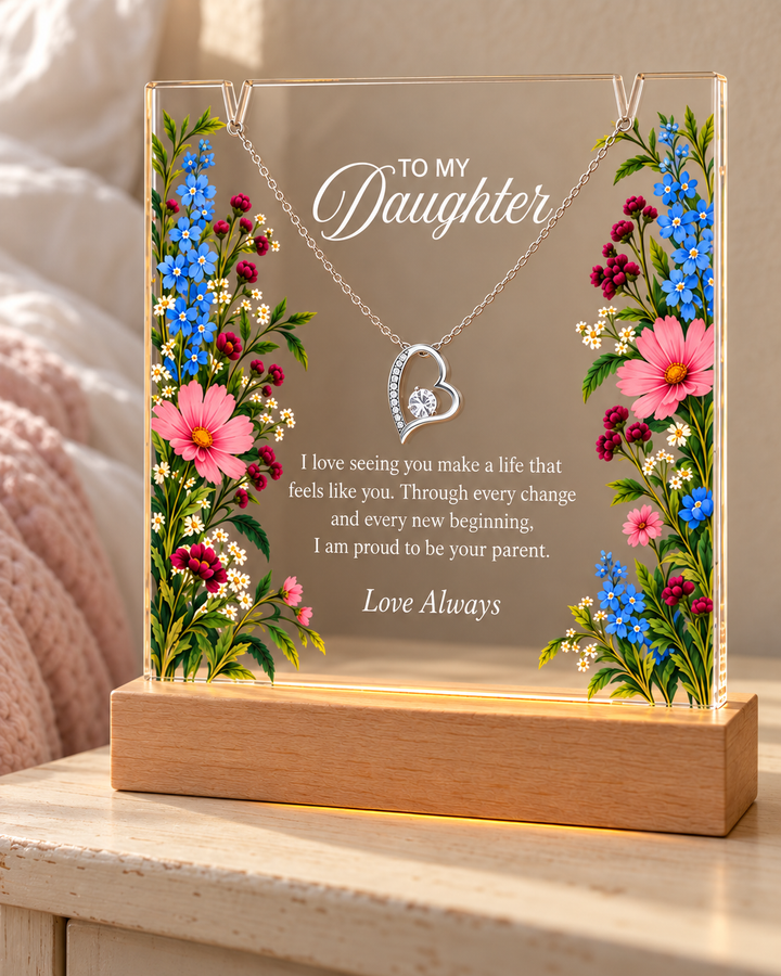 To My Daughter floral acrylic keepsake with a silver-tone heart necklace on a wooden light base beside a bed.