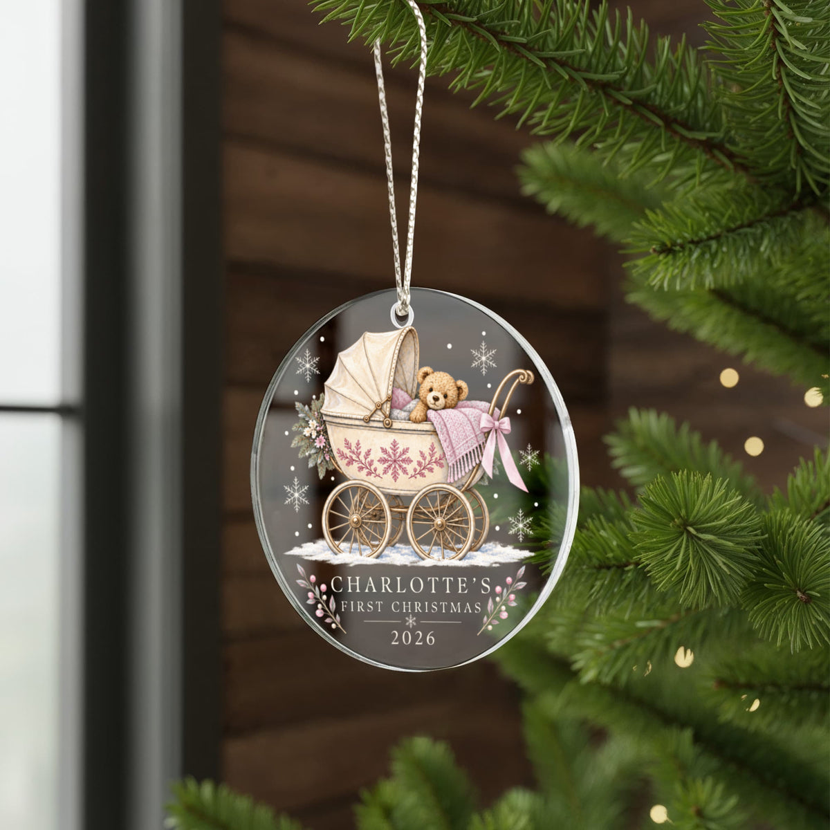Pink teddy pram first Christmas ornament hanging beside evergreen branches by a window.