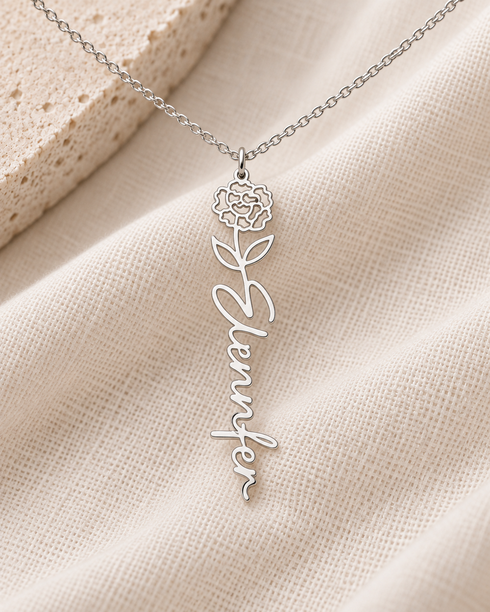Silver-tone birth flower and vertical name necklace on ivory fabric.