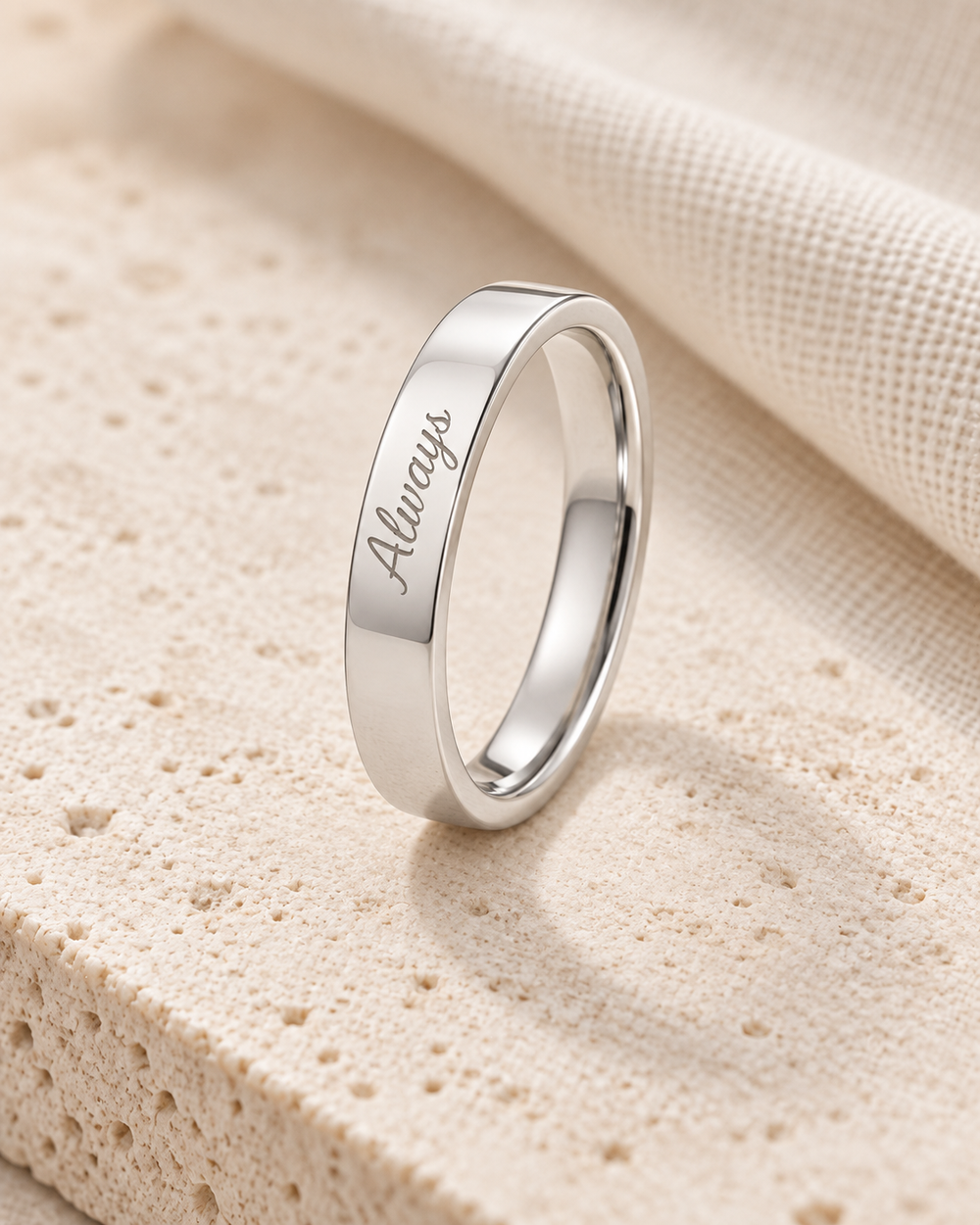 Silver-tone engraved ring standing on pale stone beside ivory fabric.