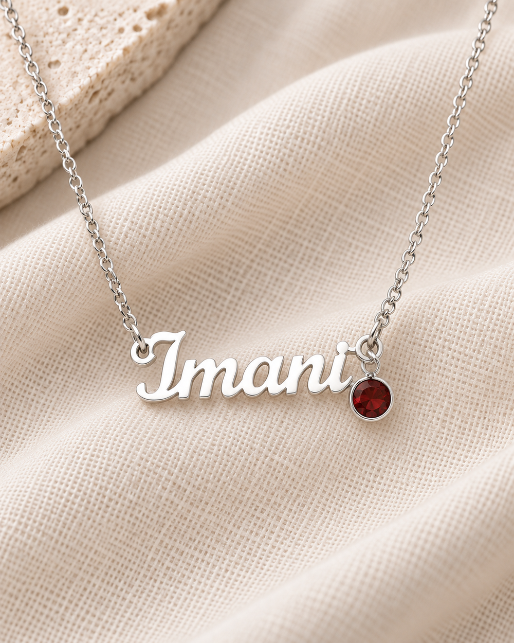 Silver-tone Imani script name necklace with a red birthstone on ivory fabric.