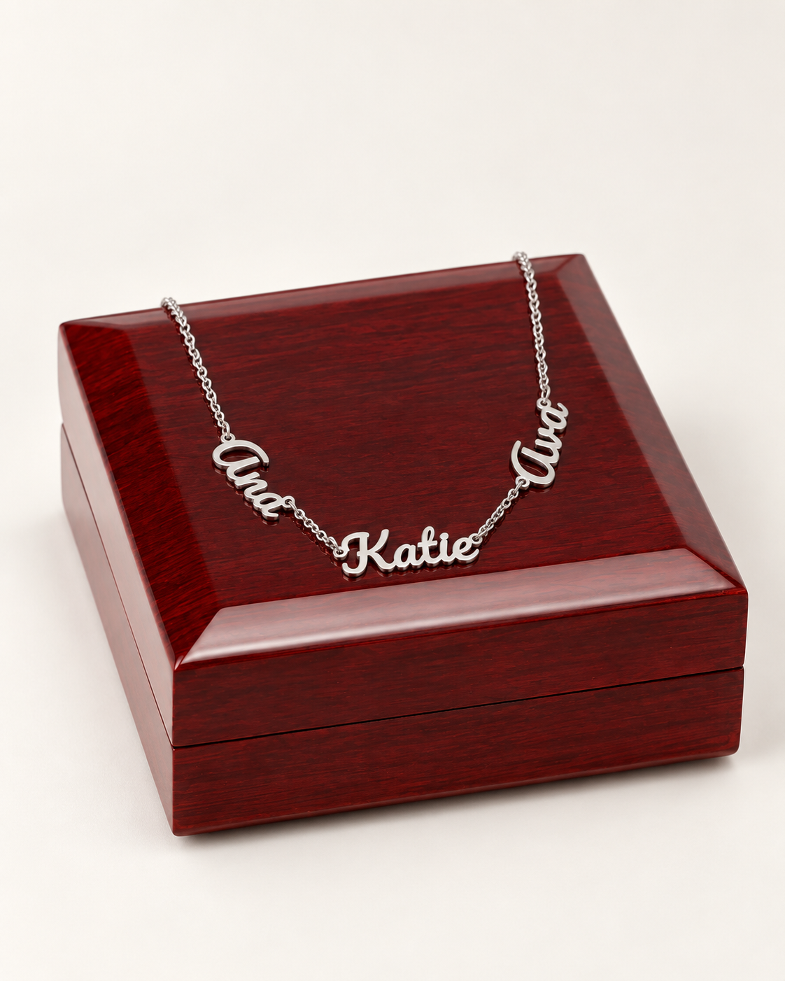 Silver-tone three-name necklace draped across a wooden gift box on ivory.